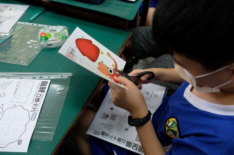國小小朋友右手拿剪刀左手握著橘色魷魚圖案紙卡低頭專注地製作卡片