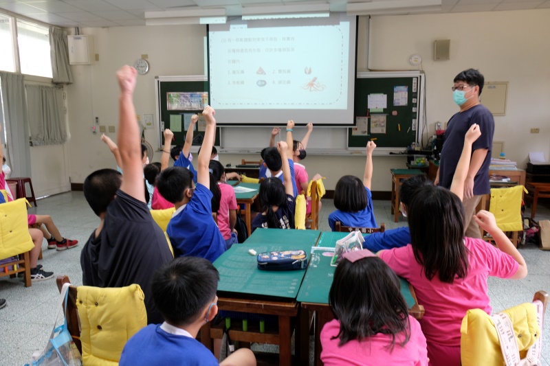 教師使用簡報為國小學童上課學生分組聽課並踴躍舉手等待老師點名發言(點擊放大)
